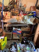 Photo showing large assortment of hand tools, cords, wood crates, cans, and miscellaneous workshop hardware items stacked and stored in a basement area.