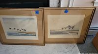 Two framed prints leaning against a wall on a marble floor. Both feature birds flying over water. Both have beige mats and gold-tone frames with visible wear. Blue tags labeled 44 and 84 on frames.