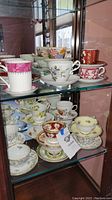Multiple china teacups and saucers arranged on glass shelves featuring various floral and ornate designs in colors such as pink, red, yellow, blue, and white.