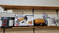 Shelf showing Rival deep fryer box, silver and black deep fryer with red controls, orange deep fryer, white food processor, and other kitchen items.