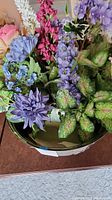 Front angle showing the metal bowl planter with detailed engraving, filled with artificial purple, blue, and pink flowers and variegated green leaves.