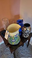 Photo showing all four items together on a wooden side table: large ceramic pitcher with floral design, cobalt blue ceramic vase, silver vase, and clear glass vase.