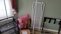 Photo showing white metal ironing board, black metal shelving units, wooden folding luggage rack, folding metal rack with fabric loops, pink bag and cardboard boxes in corner of room.