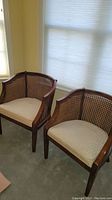 Pair of wooden side chairs with woven backs and beige cushions against wall near window.