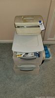 White three-drawer plastic storage unit with the top drawer open showing various maps and guidebooks inside. Beige plastic storage box sitting closed on top of the storage unit.