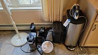 Photo showing multiple electronic and household items including an iron, trash can, shredder, coffee maker, fan, cordless phone, and vacuum cleaner on a tiled floor near a window.