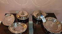 Four silverplate serving trays, two covered with dome lids and two uncovered on a small wooden table.