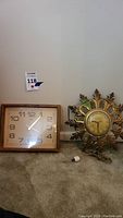 Two mid-century modern clocks placed side by side on a carpeted floor against a wall: a rectangular wood framed battery-operated clock and an ornate sunburst metal plug-in clock.