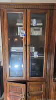Full frontal view of tall wooden display cabinet with two beveled glass doors showing multiple audio and video media devices inside including a stereo receiver and VHS player.