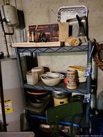 Photo shows the shelving unit mostly empty with various household items stored on it, including kitchen bowls, steins, and kitchen appliances. Shelving unit is made of dark gray plastic with four shelves.