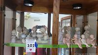 Full display cabinet shot showing multiple Precious Moments angel figurines including four sitting mud pie angels on the edge of the shelf and other standing figures behind.