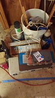 Photo showing a white bucket with cables, spray can, a metal pipe cutter, wooden items, and plastic cases likely containing sockets and bits, placed on a basement floor.