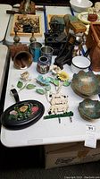 Photo of assortment on white table includes cast iron sample stove, cloisonne-style owl vase, enamelware, tin cans, metal craft flowers, painted tray, sheep wall hook, brass bell