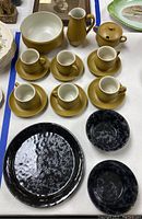 Top view of Denby mustard yellow tea set with cups, saucers, creamer, sugar bowl, serving bowl and black speckled Bennington Pottery plate and bowls.