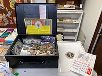 Open black Honeywell safe filled with various foreign coins, novelty currency and collectible stamps. Ameripex 86 Presidential Mint brochures placed beside the safe.