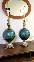 Pair of Hollywood Regency style table lamps with teal Bristol glass spheres decorated with gold floral and vine patterns, brass necks, white marble disc bases, standing on wooden furniture beneath an oval mirror.