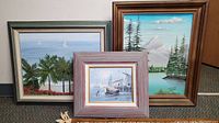 Three framed paintings grouped together showing distinct subjects: tropical beach, mountain lake, and nautical harbor.