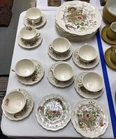 Full view of cups and saucers arranged on a table with stacked plates behind. Pattern shows flowers, birds, and fruit on off-white scalloped dishes.