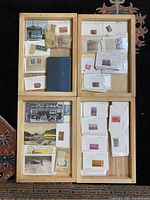 Four wooden trays arranged showing a variety of vintage stamps, postcards, and ephemera. Stamps are in envelopes and plastic sleeves, postcards depict city street and historic scenes.