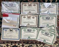 Overview photo showing multiple vintage stock certificates with cardboard backing and plastic sleeves laid out on a patterned carpet.