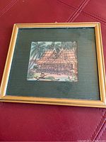 Framed Balinese art showing traditional building and figures, natural tones.