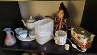 Wide shot of Asian decor items arranged on black shelf including tea set, ceramic jug, white sculptural vase, Santa figurine, ceramic teapot, small jar, and ceramic house container.