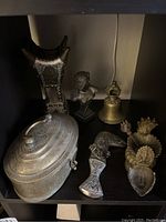 Photo showing a collection of brass and metal decor items including a covered container, decorative dagger, bust, bell, and two ornate candleholders.