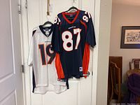 Two Denver Broncos jerseys hanging on door, one royal blue #87 and one white #19