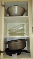 Three shelves showing large hammered metal mixing bowl, stack of three clear glass bakeware dishes, and two cast iron pans (one square skillet and one round skillet)