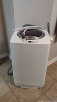 Front view of white top-loading washing machine on tiled floor with hoses draped over top