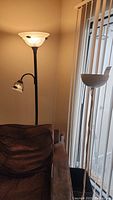 Two tall floor lamps side-by-side against wall by sliding glass door and couch, one torchiere with additional reading lamp arm, other with single bowl shade.