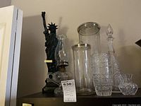 Full view of all seven items including the Liberty statue lamp, glass oil lamp, two tall glass vases, crystal decanter, tumbler and small bowl.