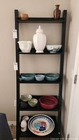 Photo of pottery items arranged on black shelving unit showing multiple pieces including white lidded urn, white textured bowl, blue-green bowls, small cream and red bowls, decorative plates and a small white teapot/pitcher.