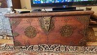 Front view of vintage wooden chest showing brass studs forming geometric and circular patterns, brass lock plate and warm brown wood surface.