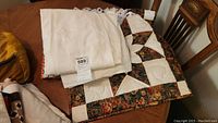 Photo showing a folded white embroidered Arabic headscarf, a cream-white Syrian tablecloth with embroidery, and an Amish quilt square piece with floral and cream quilting patterns.