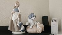 Two Nao ceramic figurines displayed side by side on a black surface against a beige wall. The larger figurine is a girl with a dog, the smaller is two puppies.