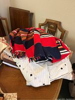 Stack of folded quilts and linens on a wooden chair and table, showing a bright red, black, white, and orange striped quilt on top with white linens beneath