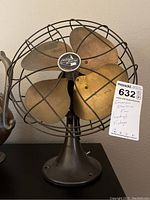 Front view of vintage Emerson Electric fan showing four large brass blades surrounded by protective wire cage, mounted on a heavy metal base with a tag noting item details.