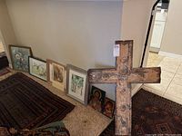 Wide view showing 9 pieces of wall art including framed prints leaning against a wall, religious icons on floor, and large rustic metal cross.