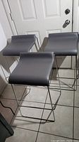 Three charcoal grey contemporary stools with chrome metal sled bases in front of a white door, showing ergonomic curved seats and backs.