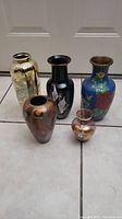 Full view of five Asian themed decorative vases arranged on floor tiles: gold brass, black with bird, blue floral enamel, copper floral, and small round gold-purple enamel.