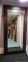 Front view of the large floor style mahogany mirror against a wall with some background reflected in the mirror.