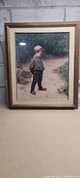 Framed print showing a boy in vintage clothing, standing near a bicycle wheel outdoors. Wooden frame with light matting visible.
