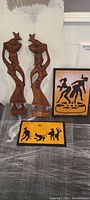 Two carved wooden human figurines and two rectangular wooden wall plaques with black carved silhouettes of dancing and horse scenes, placed on a dark surface against a wall.
