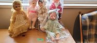 Photo of five vintage dolls grouped on a wooden table with window light. Dolls include Scottish themed doll, others in pastel dresses, some wrapped in plastic.