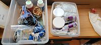 Two clear plastic bins filled with various crafting glue tools and materials including glue guns, glue sticks, bottles of adhesives, and crafting supplies arranged on a wooden surface.