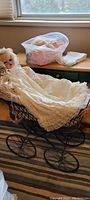 Full view of porcelain baby doll dressed in long white lace christening gown placed in vintage metal pram with large wheels. Additional baby clothes wrapped in white fabric are on the windowsill behind.
