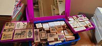 View of numerous wood-mounted rubber stamps displayed inside a blue plastic bin with a purple tray on top, showing stamps with decorative and holiday themes.