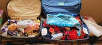 Two open vintage suitcases displaying a vintage baby doll and multiple assorted doll clothes in various colors and fabrics.