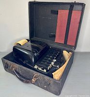 Black Stenotype Master Model 5 machine seated inside an open black carrying case with a red book strapped to the upper inside lid and worn papers inserted on the sides.